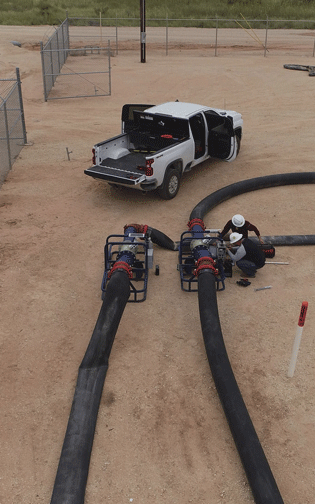 From afar, two men in hard hats leaning over equipment, black hoses, near a white truck. 