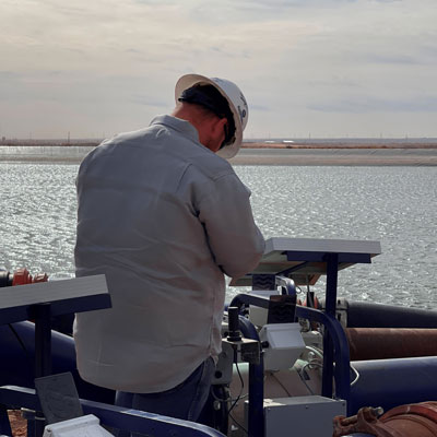 A man in a white shirt and hard hat looking down at industrial equipment, water in background.